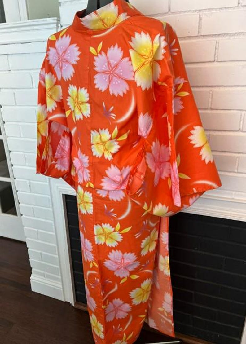 Może przedstawiać: Jasnopomarańczowe yukata z powtarzającym się kwiatowym wzorem r&oacute;żowych i ż&oacute;łtych kwiat&oacute;w wiśni. Ubi&oacute;r posiada szerokie rękawy, wysoki kołnierz oraz prosty kr&oacute;j, prezentując wyrazisty botaniczny nadruk na całej powierzchni tkaniny.