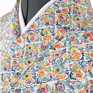 May include: Close-up of a floral patterned garment. The fabric features a white background with a repeating pattern of colorful flowers in red, yellow, blue, and green. The garment has a white collar and a V-neck design.