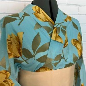 May include: A light blue kimono featuring a repeating pattern of large golden-yellow flowers and muted olive-green leaves. The garment has a wrap-style front with a wide, solid beige obi sash cinched around the waist, displayed on a mannequin.