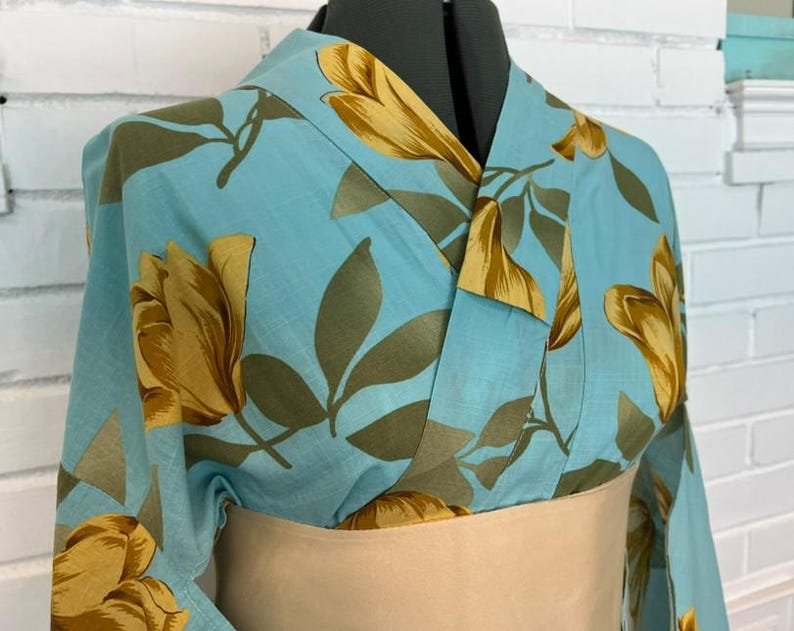 May include: A light blue kimono featuring a repeating pattern of large golden-yellow flowers and muted olive-green leaves. The garment has a wrap-style front with a wide, solid beige obi sash cinched around the waist, displayed on a mannequin.