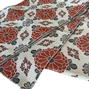 May include: A folded textile featuring a cream-coloured base with a repeating pattern of large, rust-orange chrysanthemum flowers, dark grey leaves, and small geometric diamond-shaped motifs. The fabric has a visible horizontal ribbed texture.