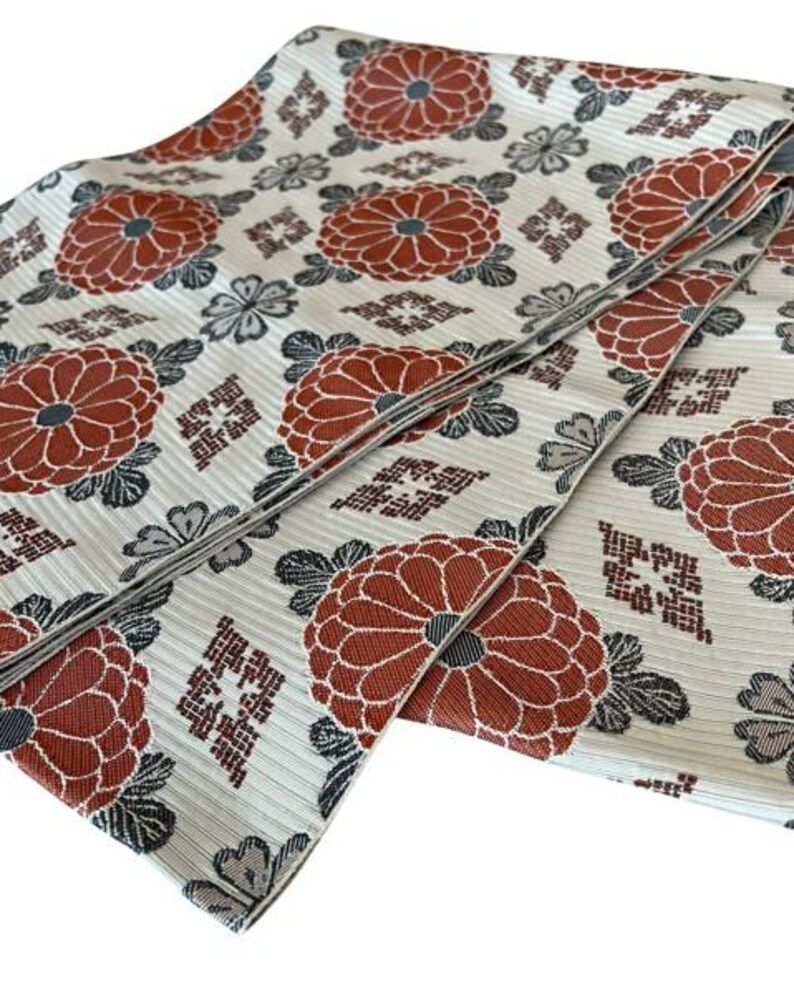 May include: A folded textile featuring a cream-coloured base with a repeating pattern of large, rust-orange chrysanthemum flowers, dark grey leaves, and small geometric diamond-shaped motifs. The fabric has a visible horizontal ribbed texture.