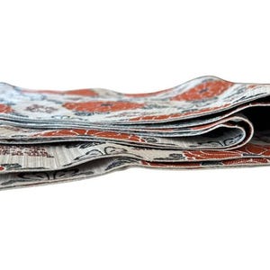 May include: A folded textile strip featuring a repeating pattern of orange flowers and dark grey foliage on a light beige background. The fabric has a visible woven texture and is layered in multiple folds, showing the edges of the material.