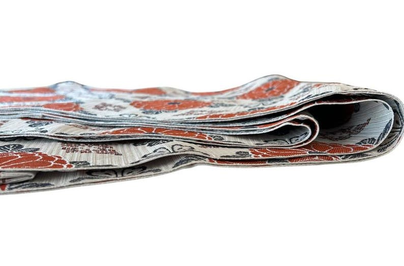 May include: A folded textile strip featuring a repeating pattern of orange flowers and dark grey foliage on a light beige background. The fabric has a visible woven texture and is layered in multiple folds, showing the edges of the material.