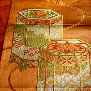 Op de afbeelding: Close-up van een decoratief textiel met twee sierlijke, zeshoekige containers. De containers hebben ingewikkelde patronen in groen, rood en goud tegen een perzikachtergrond. Het textiel bevat ook een vloeiend, oranje ontwerp met bloemaccenten.