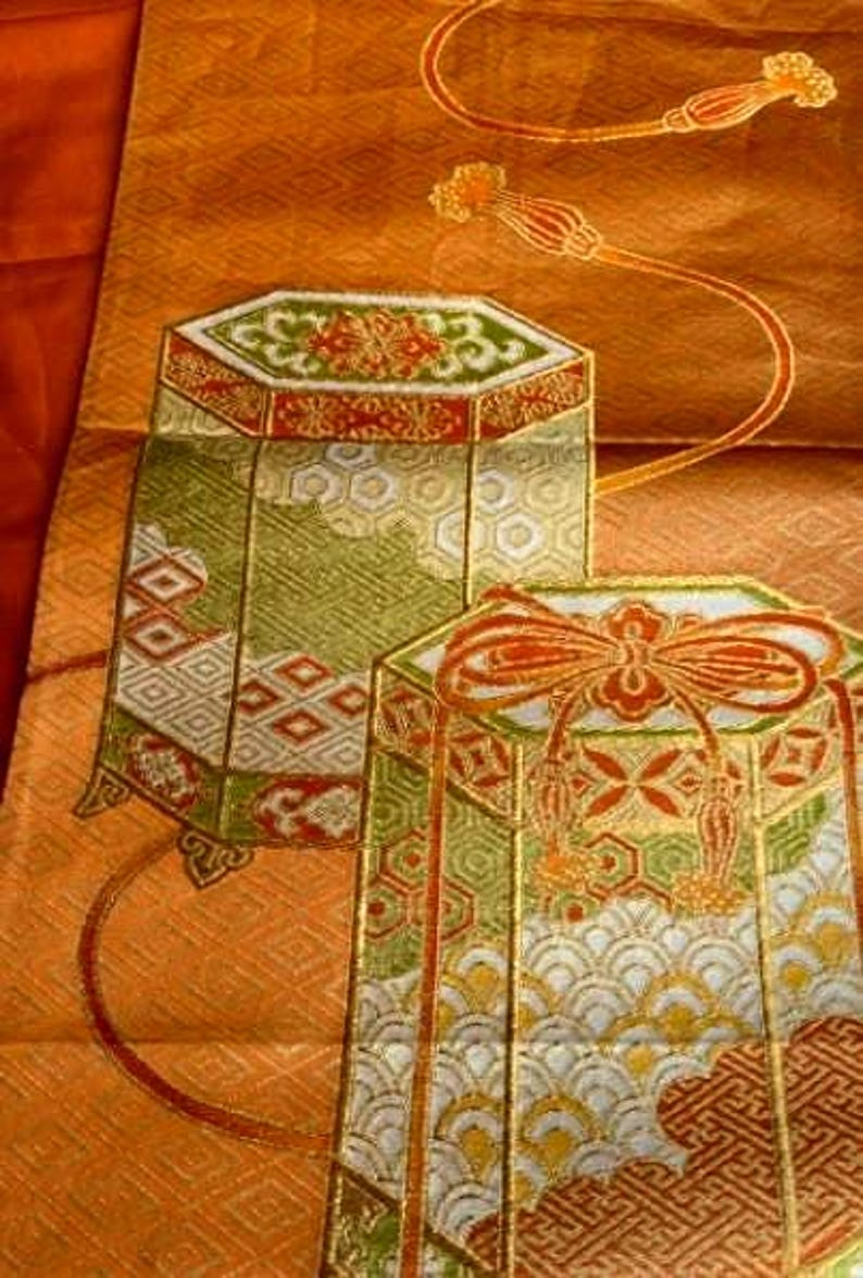 Op de afbeelding: Close-up van een decoratief textiel met twee sierlijke, zeshoekige containers. De containers hebben ingewikkelde patronen in groen, rood en goud tegen een perzikachtergrond. Het textiel bevat ook een vloeiend, oranje ontwerp met bloemaccenten.