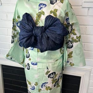 Op de afbeelding: Een licht muntgroene yukata met een bloemenpatroon van blauwe en witte ochtendgloriebloemen met groene bladeren. Het kledingstuk is in de taille getailleerd met een grote, donkerblauwe, textuurrijke obi-ceintuur die achterop in een strik is geknoopt.