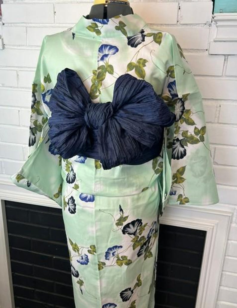 Op de afbeelding: Een licht muntgroene yukata met een bloemenpatroon van blauwe en witte ochtendgloriebloemen met groene bladeren. Het kledingstuk is in de taille getailleerd met een grote, donkerblauwe, textuurrijke obi-ceintuur die achterop in een strik is geknoopt.