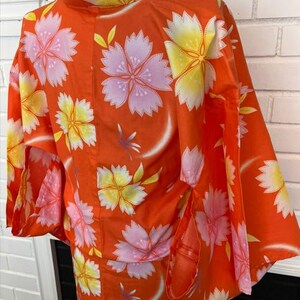 Może przedstawiać: Pomarańczowe kimono z powtarzającym się kwiatowym wzorem przedstawiającym duże r&oacute;żowe i ż&oacute;łte kwiaty. Ubi&oacute;r posiada szerokie, luźne rękawy oraz wysoki kołnierz, prezentując żywą paletę barw z delikatnymi białymi akcentami na tkaninie.