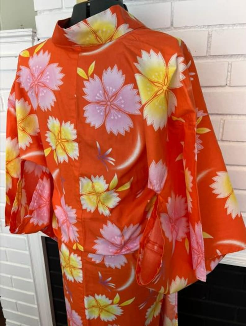 Może przedstawiać: Pomarańczowe kimono z powtarzającym się kwiatowym wzorem przedstawiającym duże r&oacute;żowe i ż&oacute;łte kwiaty. Ubi&oacute;r posiada szerokie, luźne rękawy oraz wysoki kołnierz, prezentując żywą paletę barw z delikatnymi białymi akcentami na tkaninie.
