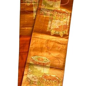 Op de afbeelding: Een lange, rechthoekige, goudoranje textiel met herhaalde geometrische patronen en gestileerde doosontwerpen in groen, goud en oranje. De stof heeft een subtiele glans en het ontwerp bevat gebogen lijnen en decoratieve elementen.