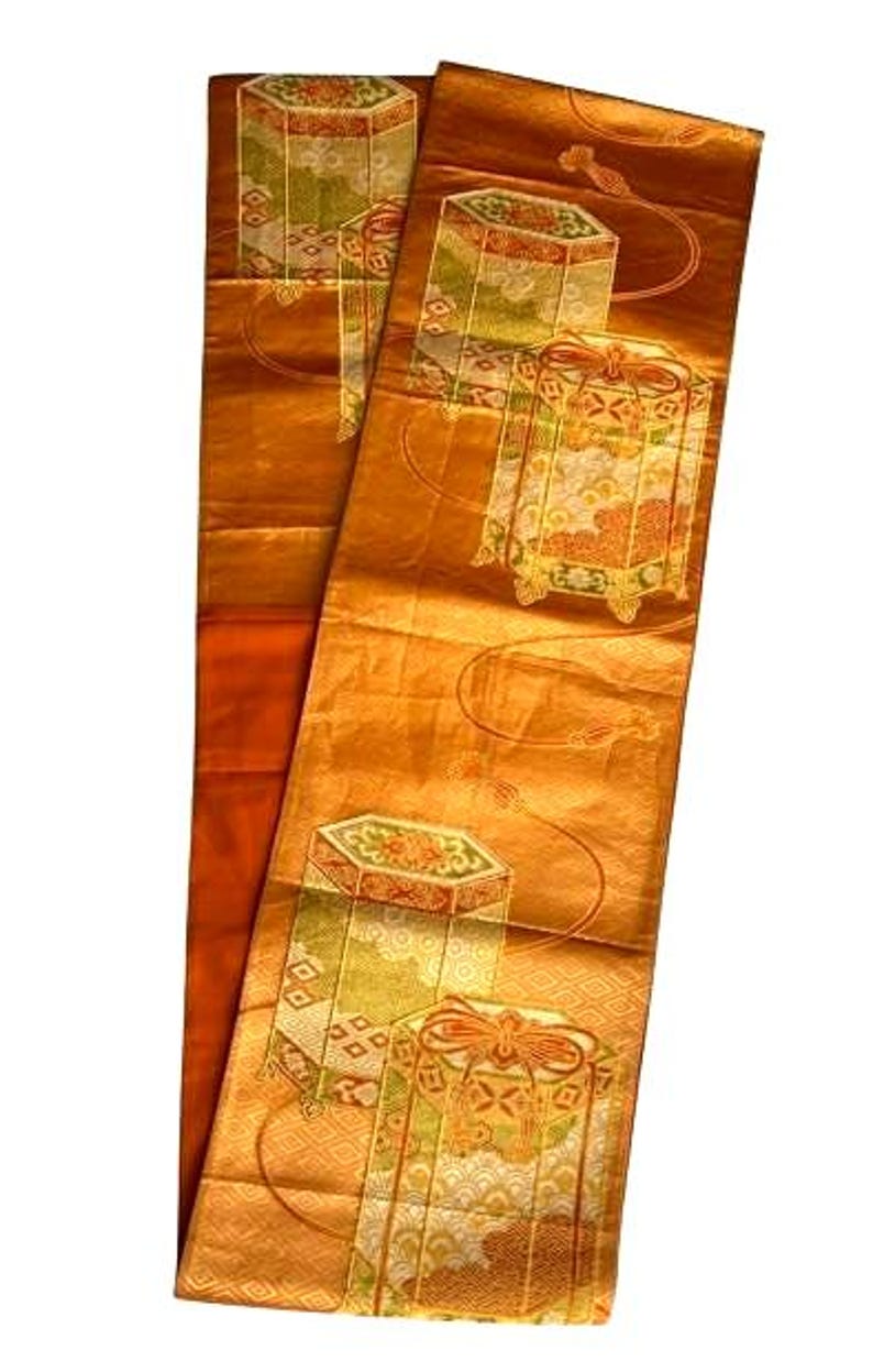 Op de afbeelding: Een lange, rechthoekige, goudoranje textiel met herhaalde geometrische patronen en gestileerde doosontwerpen in groen, goud en oranje. De stof heeft een subtiele glans en het ontwerp bevat gebogen lijnen en decoratieve elementen.