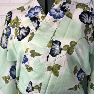 Op de afbeelding: Een lichtgroene en witte kimono met een herhalend patroon van blauwe en zwarte ochtendgloriebloemen met groene bladeren en ranken. Het kledingstuk heeft een overslagontwerp met een brede kraag en lange mouwen, getoond op een paspop.
