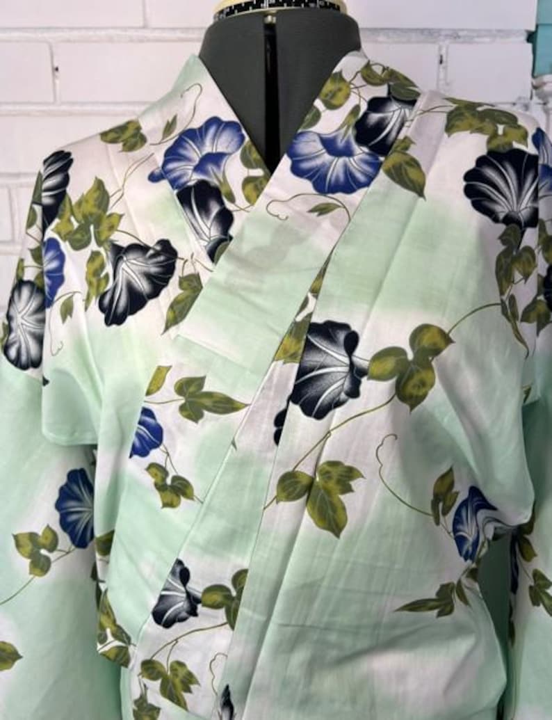 Op de afbeelding: Een lichtgroene en witte kimono met een herhalend patroon van blauwe en zwarte ochtendgloriebloemen met groene bladeren en ranken. Het kledingstuk heeft een overslagontwerp met een brede kraag en lange mouwen, getoond op een paspop.