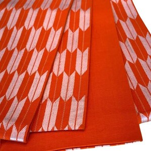 May include: A vibrant orange fabric featuring a repeating white arrow feather pattern. The textile displays a series of vertical columns of stylized arrowheads in white against a solid orange background, with one section showing solid orange.