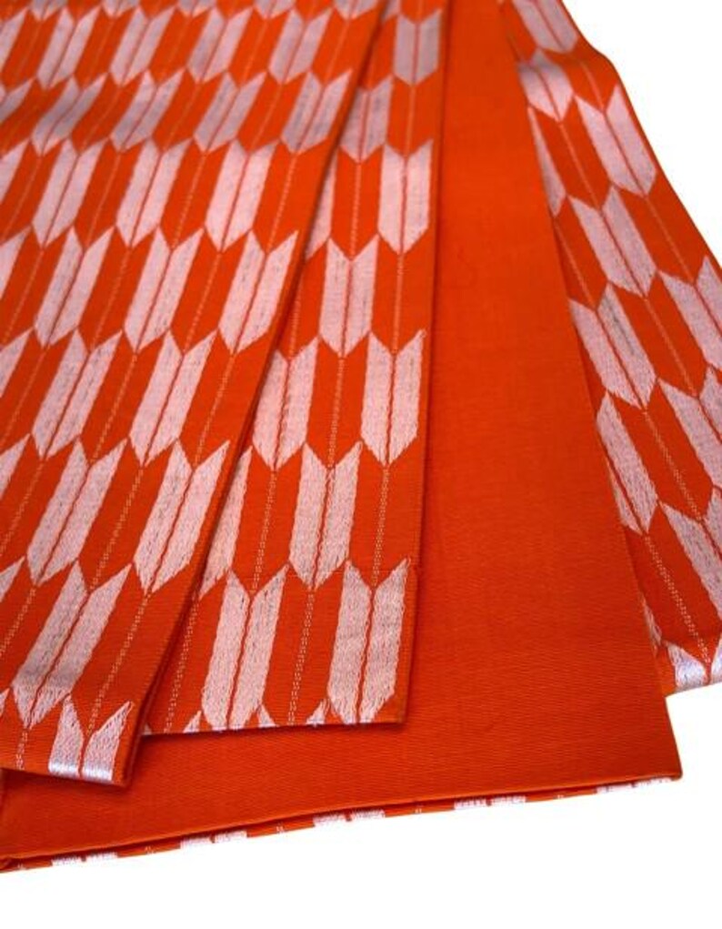 May include: A vibrant orange fabric featuring a repeating white arrow feather pattern. The textile displays a series of vertical columns of stylized arrowheads in white against a solid orange background, with one section showing solid orange.