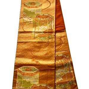Op de afbeelding: Een sierlijk, rechthoekig textiel met een herhalend patroon van geometrische vormen en gestileerde dozen in goud, oranje en groen. De stof heeft een ruitvormig patroon en een contrasterende oranje rand. Waarschijnlijk een decoratief item.