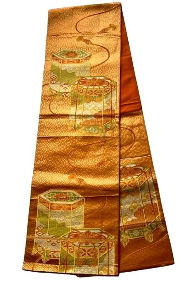 Op de afbeelding: Een sierlijk, rechthoekig textiel met een herhalend patroon van geometrische vormen en gestileerde dozen in goud, oranje en groen. De stof heeft een ruitvormig patroon en een contrasterende oranje rand. Waarschijnlijk een decoratief item.