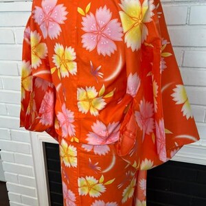 Może przedstawiać: Jaskrawopomarańczowe kimono z powtarzającym się kwiatowym wzorem w odcieniach delikatnego r&oacute;żu i jasnej ż&oacute;łci. Ubi&oacute;r posiada szerokie, luźne rękawy oraz wysoki kołnierz, prezentując żywy i kolorowy wz&oacute;r na całej powierzchni tkaniny.