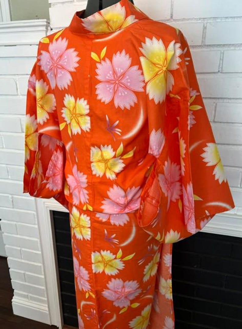 Może przedstawiać: Jaskrawopomarańczowe kimono z powtarzającym się kwiatowym wzorem w odcieniach delikatnego r&oacute;żu i jasnej ż&oacute;łci. Ubi&oacute;r posiada szerokie, luźne rękawy oraz wysoki kołnierz, prezentując żywy i kolorowy wz&oacute;r na całej powierzchni tkaniny.