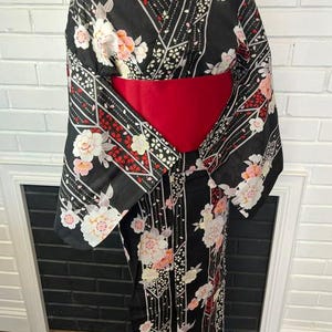 May include: A black kimono featuring a floral pattern with white and pink blossoms, accented by geometric lines and red details. The garment is cinched at the waist with a wide, solid red obi sash, creating a structured silhouette on the display form.