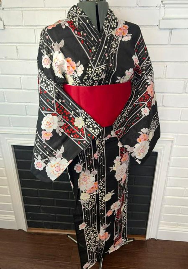 May include: A black kimono featuring a floral pattern with white and pink blossoms, accented by geometric lines and red details. The garment is cinched at the waist with a wide, solid red obi sash, creating a structured silhouette on the display form.