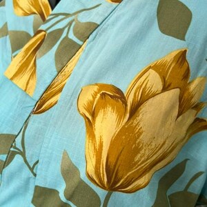 May include: A light blue robe featuring a large-scale floral pattern with golden yellow tulip-like blossoms and muted olive green leaves. The garment has a wrap-style front with a wide collar and is displayed on a mannequin torso.