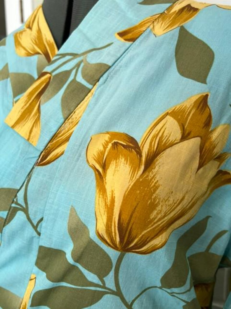 May include: A light blue robe featuring a large-scale floral pattern with golden yellow tulip-like blossoms and muted olive green leaves. The garment has a wrap-style front with a wide collar and is displayed on a mannequin torso.