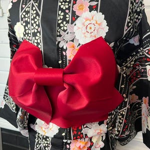 May include: A black kimono featuring a repeating pattern of white and pink floral blossoms and geometric lines, accented by a large, solid red satin bow tied at the waist. The garment has wide sleeves and a structured, smooth fabric finish.