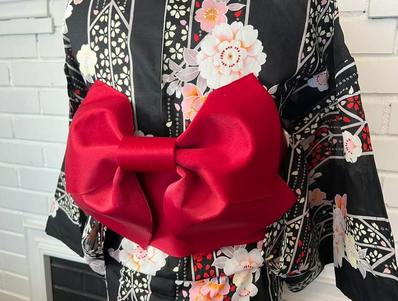 May include: A black kimono featuring a repeating pattern of white and pink floral blossoms and geometric lines, accented by a large, solid red satin bow tied at the waist. The garment has wide sleeves and a structured, smooth fabric finish.