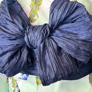 Op de afbeelding: Een grote, donker marineblauwe stoffen strik met een gekreukte textuur, gebonden rond een lichtgroen kledingstuk met een bloemenpatroon van blauwe ochtendgloriebloemen en groene bladeren. De strik is centraal geplaatst en domineert het beeld.