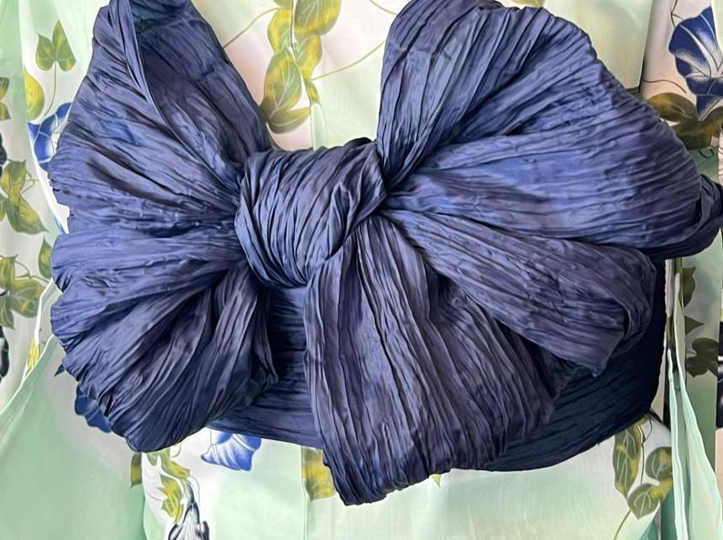 Op de afbeelding: Een grote, donker marineblauwe stoffen strik met een gekreukte textuur, gebonden rond een lichtgroen kledingstuk met een bloemenpatroon van blauwe ochtendgloriebloemen en groene bladeren. De strik is centraal geplaatst en domineert het beeld.