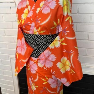 Może przedstawiać: Jaskrawopomarańczowe kimono z powtarzającym się wzorem dużych r&oacute;żowych i ż&oacute;łtych kwiat&oacute;w wiśni oraz małych ważek. Ubi&oacute;r jest ściągnięty w talii szerokim, czarnym pasem obi z białym, misternym geometrycznym wzorem przypominającym kratkę.