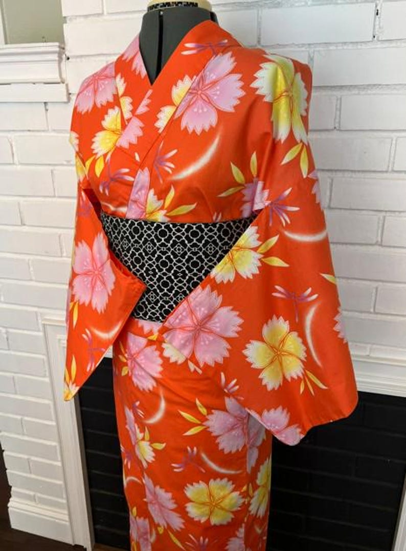 Może przedstawiać: Jaskrawopomarańczowe kimono z powtarzającym się wzorem dużych r&oacute;żowych i ż&oacute;łtych kwiat&oacute;w wiśni oraz małych ważek. Ubi&oacute;r jest ściągnięty w talii szerokim, czarnym pasem obi z białym, misternym geometrycznym wzorem przypominającym kratkę.