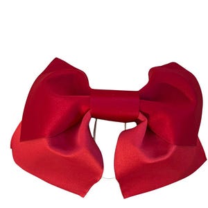 May include: A deep red hair bow featuring a smooth, satin-like finish with wide, structured loops and a central knot. The accessory has a symmetrical design with two large, folded wings extending outward and a clear elastic band visible at the back.