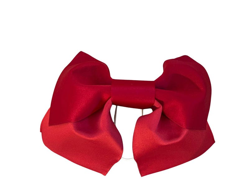 May include: A deep red hair bow featuring a smooth, satin-like finish with wide, structured loops and a central knot. The accessory has a symmetrical design with two large, folded wings extending outward and a clear elastic band visible at the back.