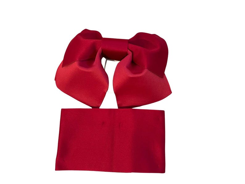 May include: A deep red satin hair accessory consisting of a large, structured bow with two wide, folded loops and a separate, rectangular fabric band positioned directly below the bow, both featuring a smooth, reflective, and uniform finish.