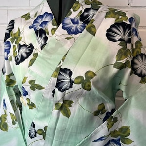 Op de afbeelding: Een kimono met een licht mintgroene achtergrond en een herhalend patroon van blauwe en zwarte ochtendgloriebloemen en groene bladeren. Het kledingstuk heeft een overslagontwerp met wijde mouwen en een gestructureerde kraag met bloemenprint.