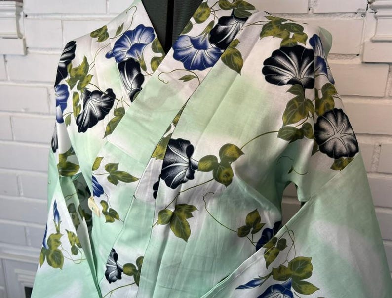 Op de afbeelding: Een kimono met een licht mintgroene achtergrond en een herhalend patroon van blauwe en zwarte ochtendgloriebloemen en groene bladeren. Het kledingstuk heeft een overslagontwerp met wijde mouwen en een gestructureerde kraag met bloemenprint.