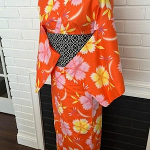 Może przedstawiać: Jasnopomarańczowe kimono z powtarzającym się wzorem dużych r&oacute;żowych i ż&oacute;łtych kwiat&oacute;w wiśni. Ubi&oacute;r posiada szerokie rękawy oraz kontrastujący czarno-biały pas obi z geometrycznym wzorem owinięty wok&oacute;ł talii.