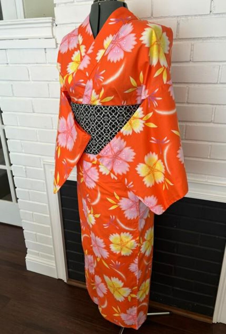Może przedstawiać: Jasnopomarańczowe kimono z powtarzającym się wzorem dużych r&oacute;żowych i ż&oacute;łtych kwiat&oacute;w wiśni. Ubi&oacute;r posiada szerokie rękawy oraz kontrastujący czarno-biały pas obi z geometrycznym wzorem owinięty wok&oacute;ł talii.