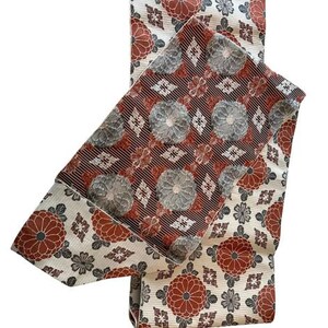 May include: A folded textile sash featuring a cream-coloured base with a repeating pattern of rust-orange and grey floral motifs. The centre section displays a contrasting dark brown background with a dense pattern of grey and rust-orange circular flowers.