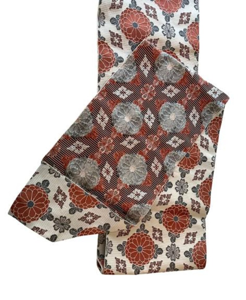 May include: A folded textile sash featuring a cream-coloured base with a repeating pattern of rust-orange and grey floral motifs. The centre section displays a contrasting dark brown background with a dense pattern of grey and rust-orange circular flowers.