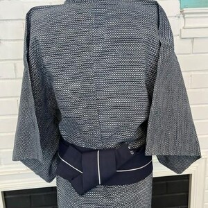 Może przedstawiać: Kimono w ciemnoniebiesko-biały wz&oacute;r wykonane z teksturowanej tkaniny z powtarzającym się geometrycznym motywem kropek. Odzież jest przewiązana w talii jednolitym, ciemnogranatowym pasem obi z pojedynczym, cienkim, białym pionowym paskiem.