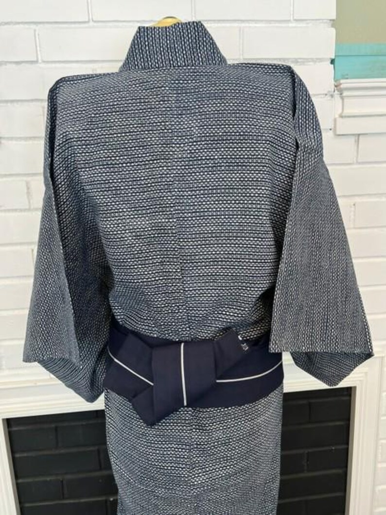 Może przedstawiać: Kimono w ciemnoniebiesko-biały wz&oacute;r wykonane z teksturowanej tkaniny z powtarzającym się geometrycznym motywem kropek. Odzież jest przewiązana w talii jednolitym, ciemnogranatowym pasem obi z pojedynczym, cienkim, białym pionowym paskiem.