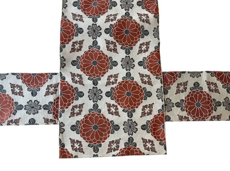 May include: A textile fabric featuring a repeating pattern of large, circular, rust-coloured floral motifs interspersed with smaller, grey, four-petaled blossoms and geometric diamond shapes on a textured, off-white background.