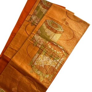 Op de afbeelding: Een oranje textiel met ingewikkelde patronen en ontwerpen. De stof heeft geometrische vormen, bloemmotieven en decoratieve doosachtige illustraties in tinten groen, rood en goud. Het textiel is gevouwen en onthult lagen van het patroonmateriaal.