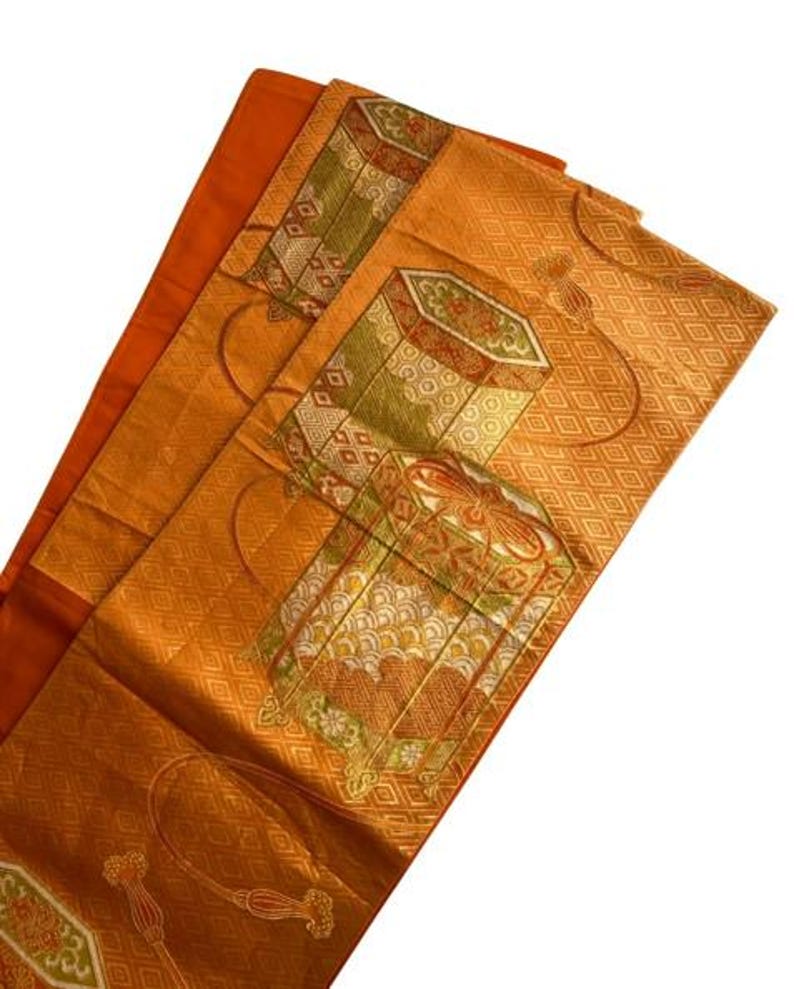Op de afbeelding: Een oranje textiel met ingewikkelde patronen en ontwerpen. De stof heeft geometrische vormen, bloemmotieven en decoratieve doosachtige illustraties in tinten groen, rood en goud. Het textiel is gevouwen en onthult lagen van het patroonmateriaal.