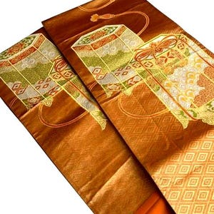 Op de afbeelding: Close-up van een gevouwen, sierlijk textiel met een rijke, bronsbruine tint. De stof heeft ingewikkelde geometrische en bloemmotieven in goud, groen en rood. Het ontwerp bevat gestileerde doosachtige motieven en decoratieve randen.