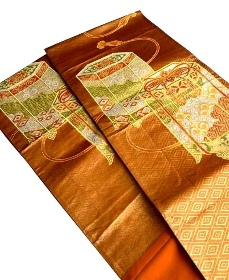 Op de afbeelding: Close-up van een gevouwen, sierlijk textiel met een rijke, bronsbruine tint. De stof heeft ingewikkelde geometrische en bloemmotieven in goud, groen en rood. Het ontwerp bevat gestileerde doosachtige motieven en decoratieve randen.